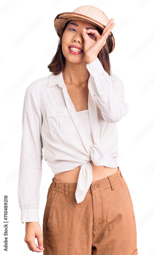 Young beautiful chinese girl wearing explorer hat smiling happy doing ok sign with hand on eye looking through fingers Young beautiful chinese girl wearing explorer hat smiling happy doing ok sign with hand on eye looking through fingers