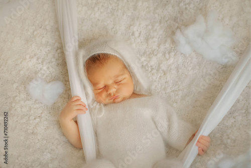 A newborn baby sleeps sweetly. Baby in a white fluffy bodysuit. Bunny boy. The boy is lying on a white blanket. The baby is on a swing. Son. Newborn photo session. Baby in the clouds. Dreams. Tenderne photo