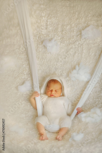 A newborn baby sleeps sweetly. Baby in a white fluffy bodysuit. Bunny boy. The boy is lying on a white blanket. The baby is on a swing. Son. Newborn photo session. Baby in the clouds. Dreams. Tenderne photo