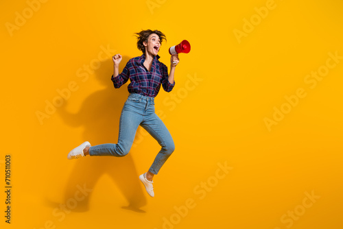 Full size photo of attractive eccentric girl jumping scream in megaphone look empty space support team isolated on yellow color background