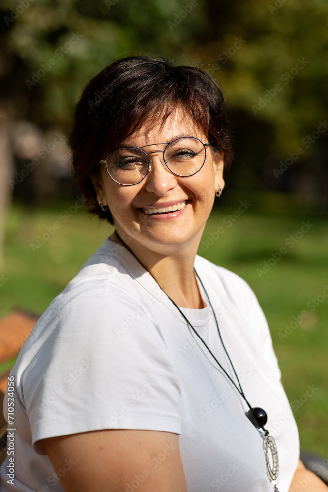 A beautiful middle-aged woman with glasses and short dark hair is sitting on a bench A beautiful middle-aged woman with glasses and short dark hair is sitting on a bench