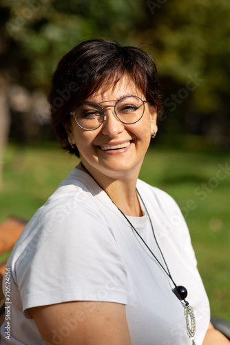 A beautiful middle-aged woman with glasses and short dark hair is sitting on a bench