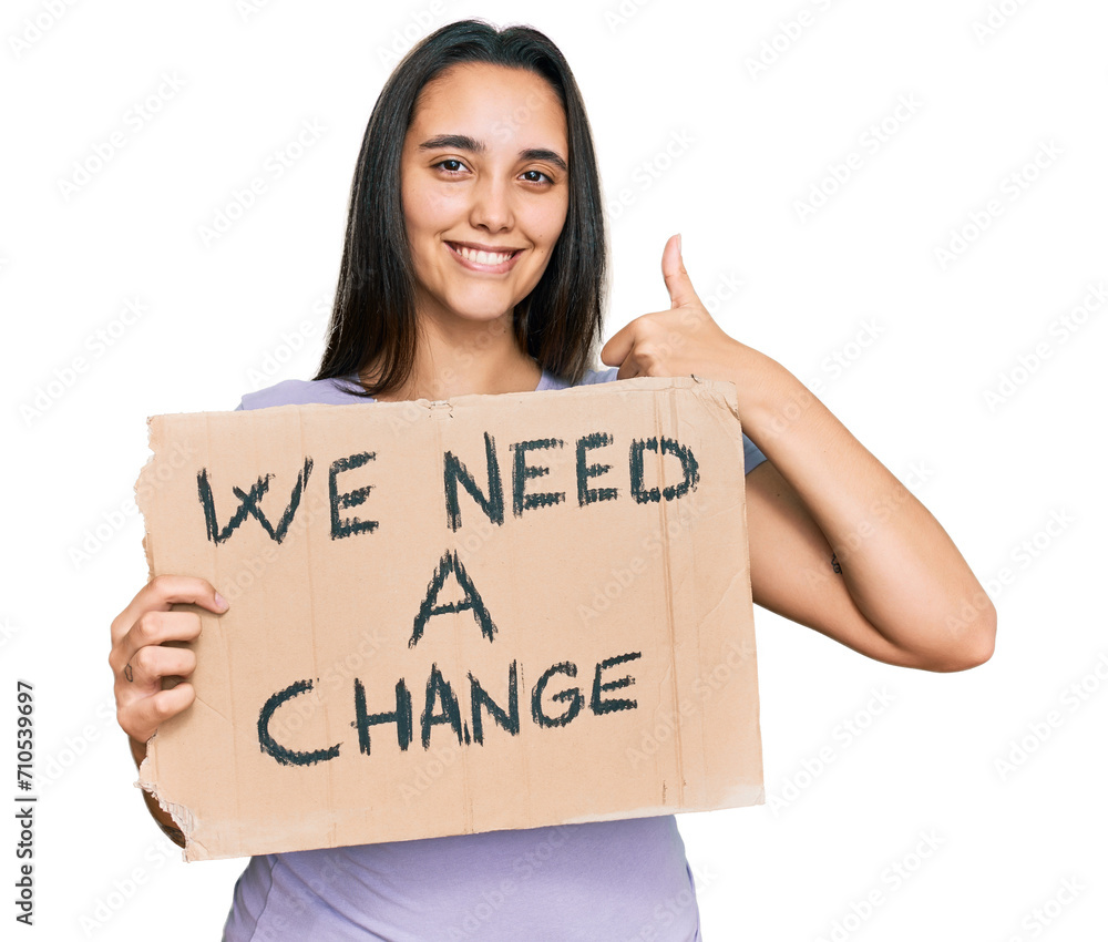 Young hispanic woman holding we need a change banner smiling happy and positive, thumb up doing excellent and approval sign Young hispanic woman holding we need a change banner smiling happy and positive, thumb up doing excellent and approval sign