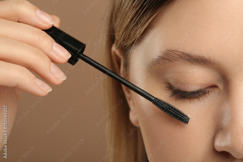 Woman applying mascara onto eyelashes against light brown background, closeup