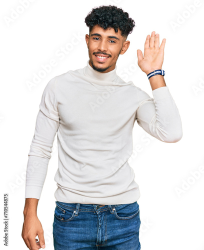 Young arab man wearing casual winter sweater waiving saying hello happy and smiling, friendly welcome gesture