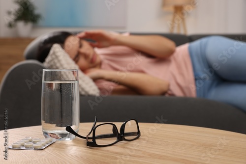 Sad woman suffering from headache indoors, focus on glass of water, pills and eyeglasses