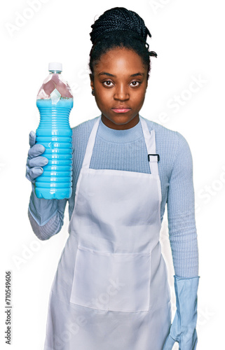 Young african american woman wearing apron holding detergent bottle thinking attitude and sober expression looking self confident