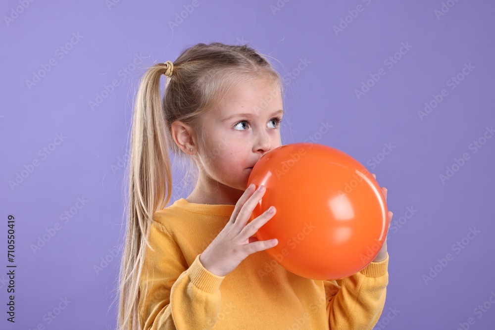 Cute little girl inflating orange balloon on violet background Cute little girl inflating orange balloon on violet background