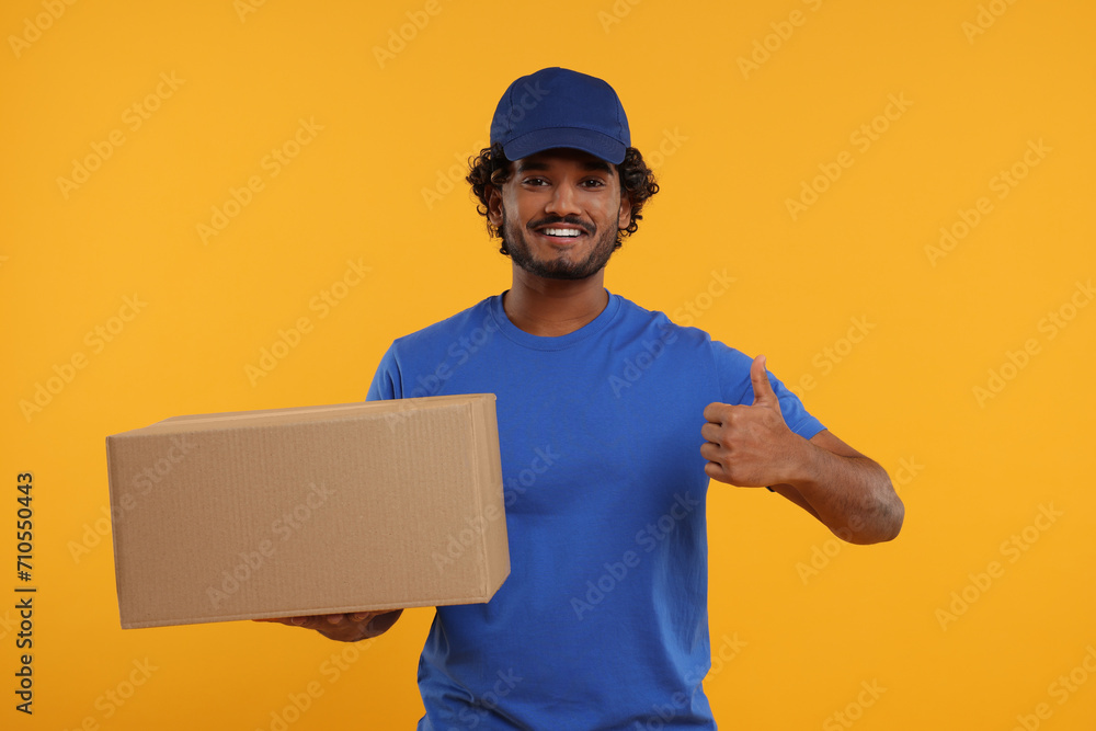 Happy young courier with parcel showing thumb up on orange background