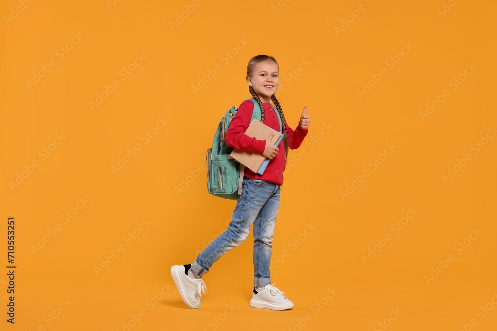 Happy schoolgirl with backpack and books showing thumb up gesture on orange background Happy schoolgirl with backpack and books showing thumb up gesture on orange background