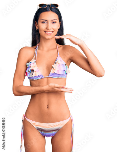 Young beautiful latin girl wearing bikini and sunglasses gesturing with hands showing big and large size sign, measure symbol. smiling looking at the camera. measuring concept.