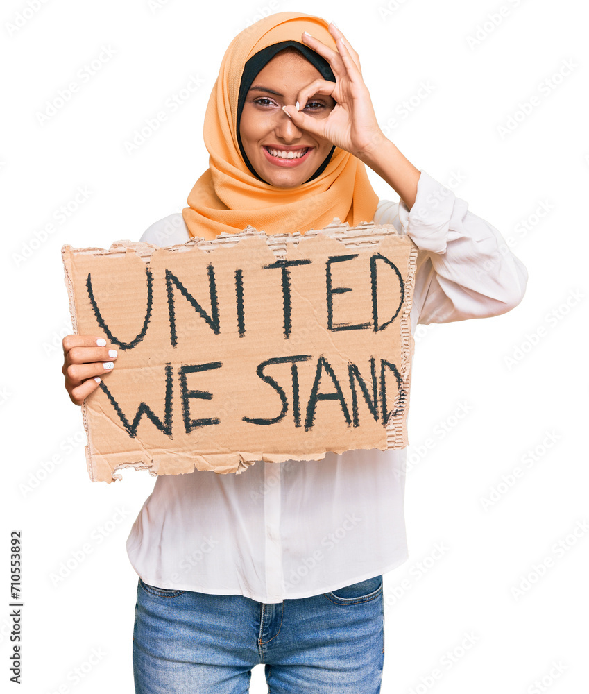 Young brunette arab woman holding united we stand banner smiling happy doing ok sign with hand on eye looking through fingers Young brunette arab woman holding united we stand banner smiling happy doing ok sign with hand on eye looking through fingers
