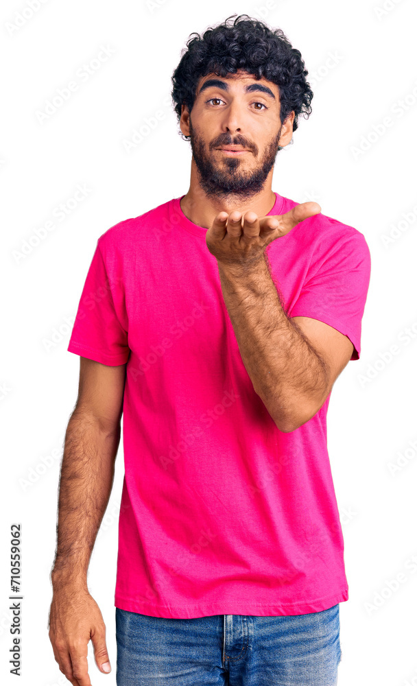 Handsome young man with curly hair and bear wearing casual pink tshirt looking at the camera blowing a kiss with hand on air being lovely and sexy. love expression. Handsome young man with curly hair and bear wearing casual pink tshirt looking at the camera blowing a kiss with hand on air being lovely and sexy. love expression.