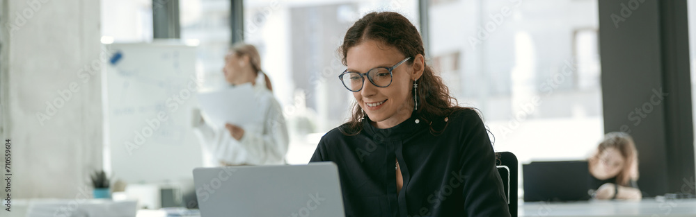 Smiling stylish businesswoman working on laptop sitting in modern office on colleagues background Smiling stylish businesswoman working on laptop sitting in modern office on colleagues background