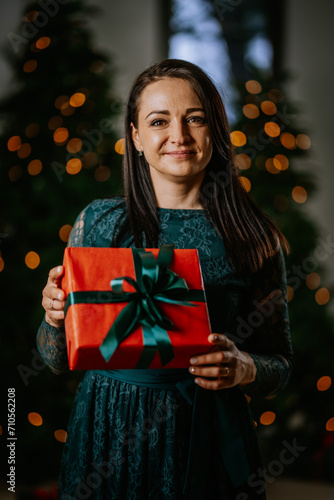 woman holding a red gift with a green ribbon. She's smiling, standing in front of a Christmas tree with lights...