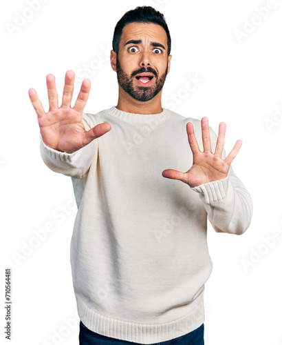 Young hispanic man with beard wearing casual white sweater afraid and terrified with fear expression stop gesture with hands, shouting in shock. panic concept.