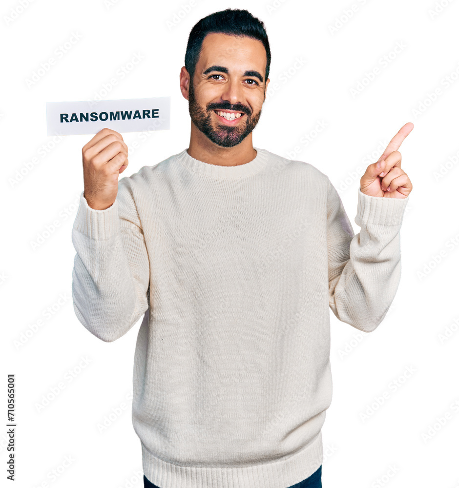 Young hispanic man with beard holding ransomware banner smiling happy pointing with hand and finger to the side Young hispanic man with beard holding ransomware banner smiling happy pointing with hand and finger to the side