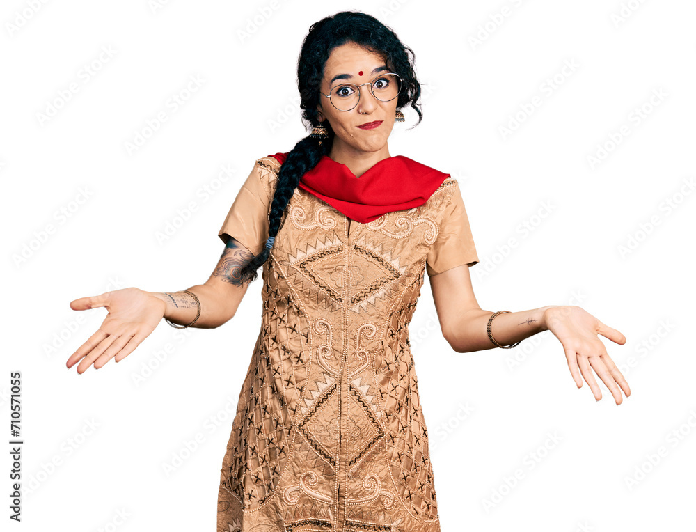 Young indian woman wearing traditional dress and glasses clueless and confused expression with arms and hands raised. doubt concept. Young indian woman wearing traditional dress and glasses clueless and confused expression with arms and hands raised. doubt concept.