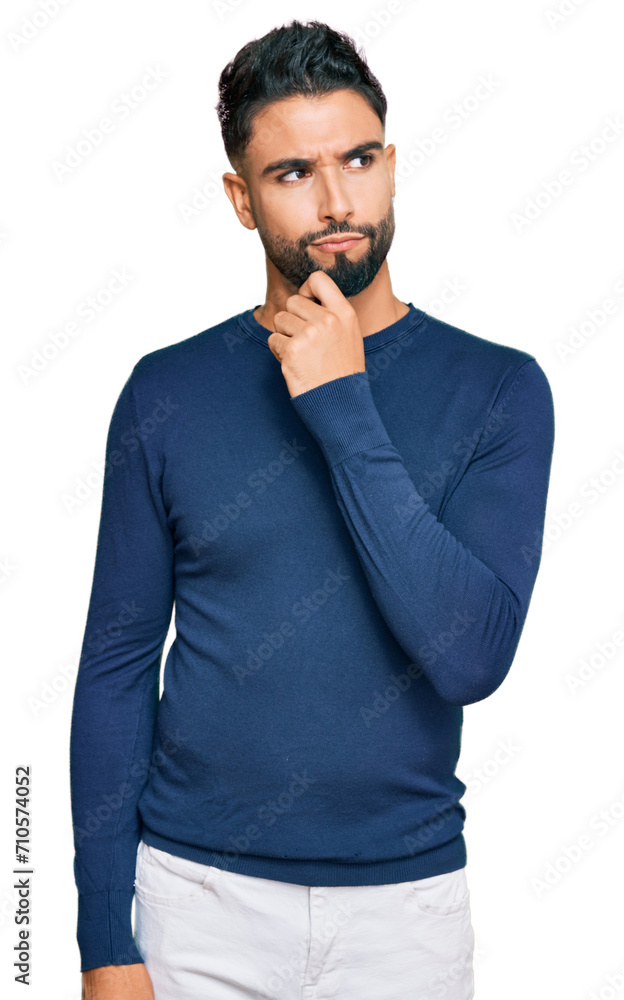 Young man with beard wearing casual blue winter sweater serious face thinking about question with hand on chin, thoughtful about confusing idea Young man with beard wearing casual blue winter sweater serious face thinking about question with hand on chin, thoughtful about confusing idea