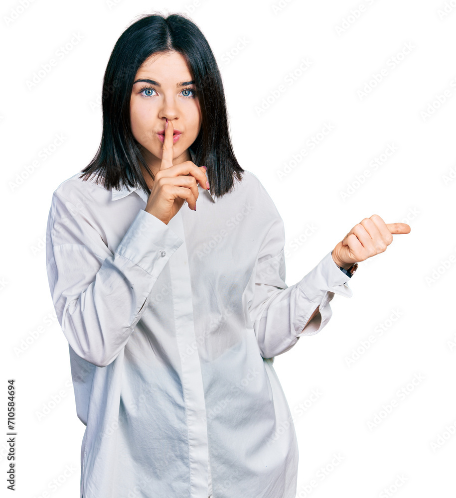 Young brunette woman with blue eyes wearing oversize white shirt asking to be quiet with finger on lips pointing with hand to the side. silence and secret concept. Young brunette woman with blue eyes wearing oversize white shirt asking to be quiet with finger on lips pointing with hand to the side. silence and secret concept.