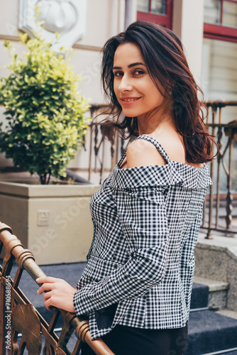 A smiling woman with shoulder-length hair stands on a city street, her hand resting on a decorative railing as she looks back over her shoulder, wearing a chic off-shoulder gingham top