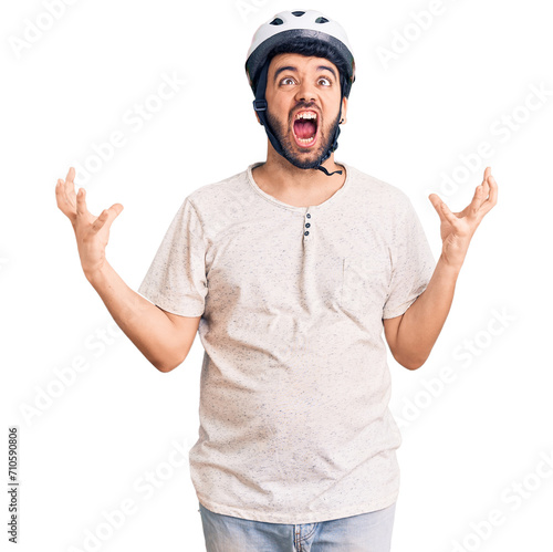 Young hispanic man wearing bike helmet crazy and mad shouting and yelling with aggressive expression and arms raised. frustration concept.