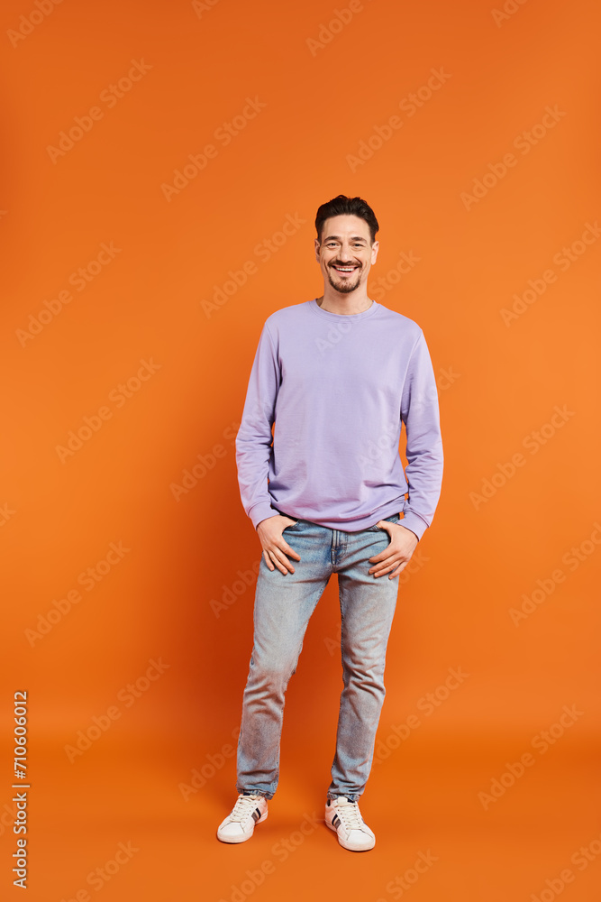 full length of cheerful and bearded man in purple sweater looking at camera on orange background full length of cheerful and bearded man in purple sweater looking at camera on orange background