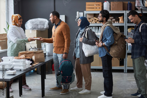 Full length side view of group of Middle Eastern people standing in line at refugee help center for free hot meals and supplies photo