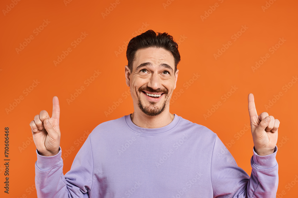cheerful man in purple sweatshirt pointing up with fingers on orange background, presenting cheerful man in purple sweatshirt pointing up with fingers on orange background, presenting