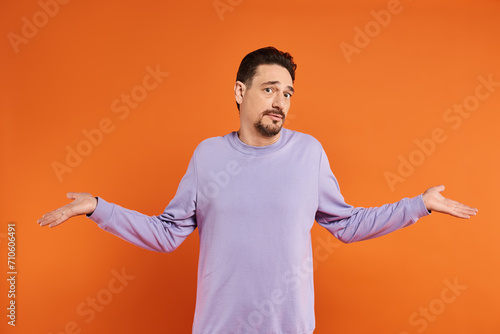 confused bearded man in purple sweater showing shrug gesture with his hands on orange background photo