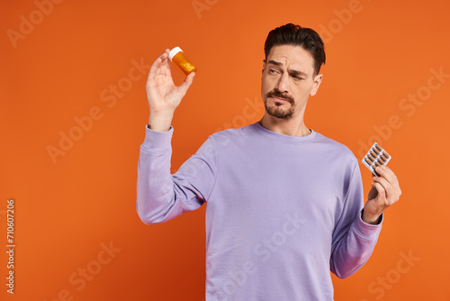 bearded man in purple sweatshirt holding bottle with pills and blister pack on orange background photo