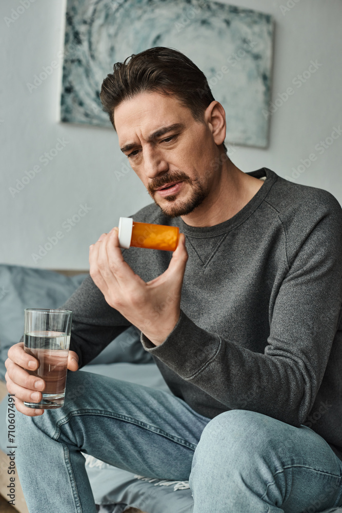 Fototapeta premium bearded man in grey jumper frowning and holding glass of water while looking at bottle with pills