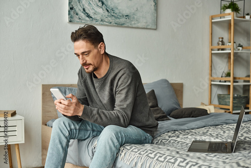 bearded man texting on mobile phone while working remotely from home near laptop on bed, freelance photo