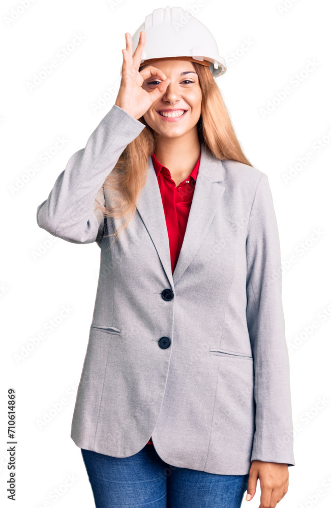 Beautiful young woman wearing architect hardhat doing ok gesture with hand smiling, eye looking through fingers with happy face.