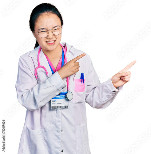Young chinese woman wearing doctor uniform and stethoscope pointing aside worried and nervous with both hands  concerned and surprised expression