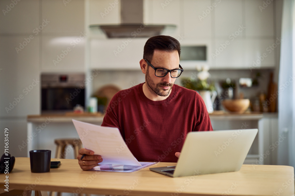 A focused freelance man with glasses using a laptop for his remote work and holding a document. A focused freelance man with glasses using a laptop for his remote work and holding a document.