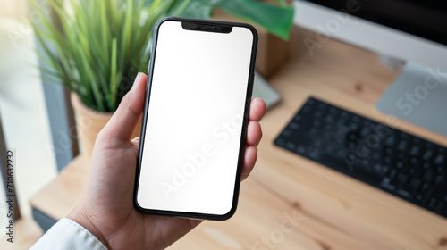  A close up of a phone with a plant and desk in the background. In front of a keyboard. Blank white screen mobile device. Insert your own screen mockup. For app design and marketing.