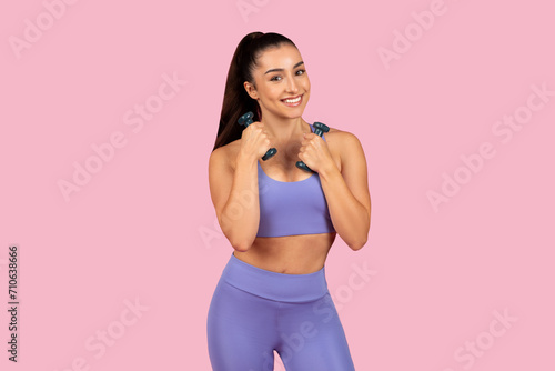 Smiling caucasian lady in sportswear doing exercises with hand dumbbells, enjoying workout on pink background
