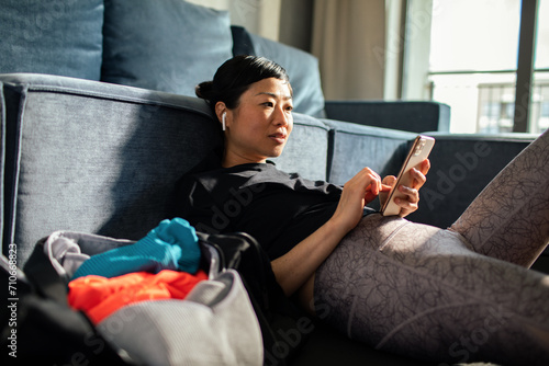 Asian woman relaxing on couch with smartphone and gym bag
