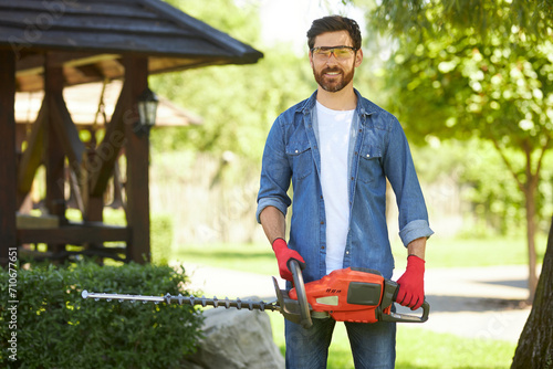 Confident gardener in protective glasses smiling, while posing with electric hedge trimmer outdoors. Portrait of happy handyman with bush cutter looking at camera, while working. Concept of lifestyle. photo