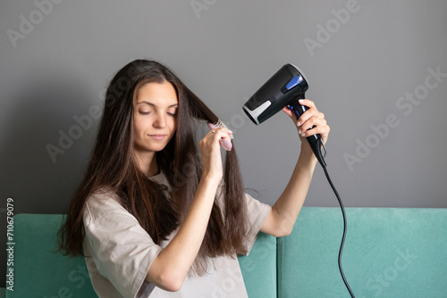 A woman comfortably styles her long dark hair using a hairdryer and round brush in a home setting, showcasing a common beauty routine.
