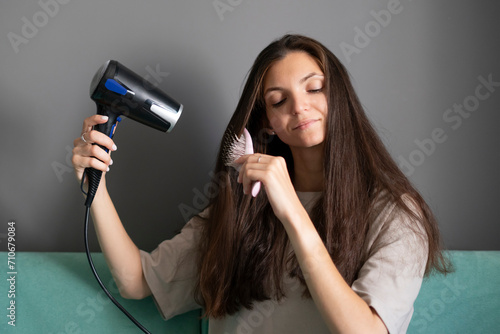 A woman comfortably styles her long dark hair using a hairdryer and round brush in a home setting, showcasing a common beauty routine.