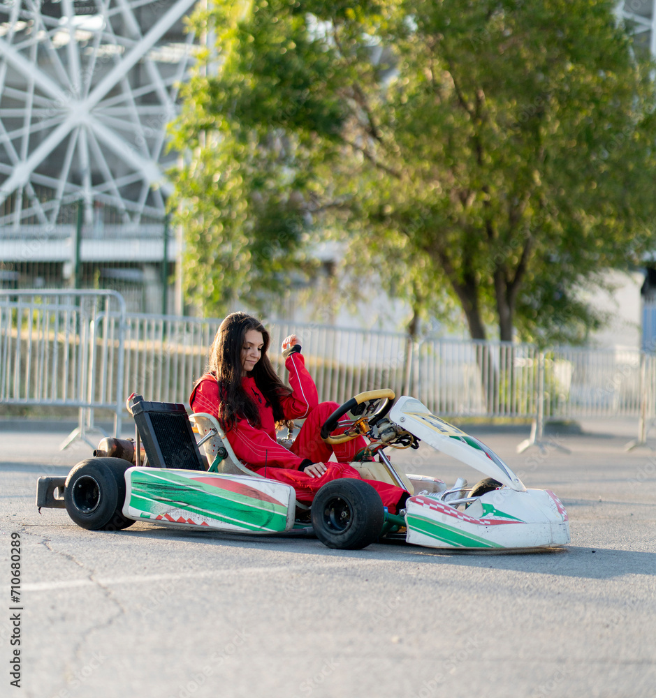 A young girl wearing a red racing suit is seated in a go-kart, ready to race on an outdoor asphalt track, showcasing determination and focus.