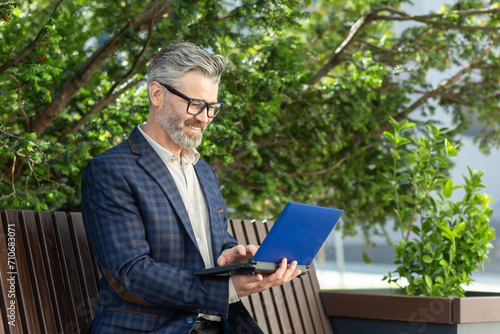 Elegant senior businessman with glasses using a digital tablet while sitting on a bench surrounded by greenery.