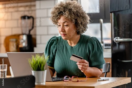 A mature woman of mixed race is making an online purchase or payment  utilizing a credit card and a laptop computer. Online banking concept