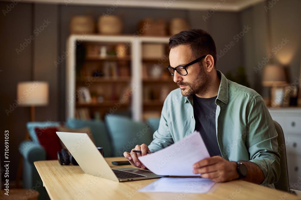 A focused adult male accountant working from home on his laptop and holding documents in his hand.