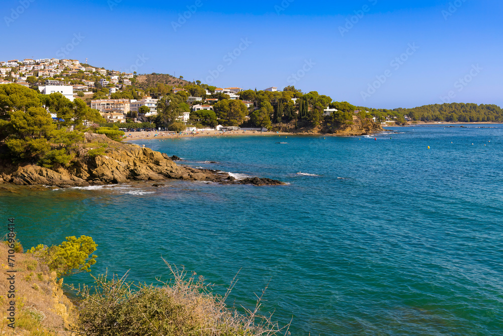 View of the coast of Colera, in Alt Empordà, with turquoise waters and Mediterranean vegetation. White houses are scattered on the hillside under a blue sky.