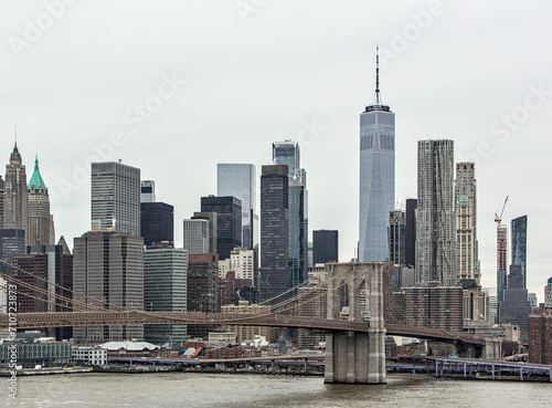 nyc skyline with brooklyn bridge and one world trade center  gray sky  cloudy overcast day  downtown view from manhattan bridge with dumbo carousel and ferry boat on hudson river