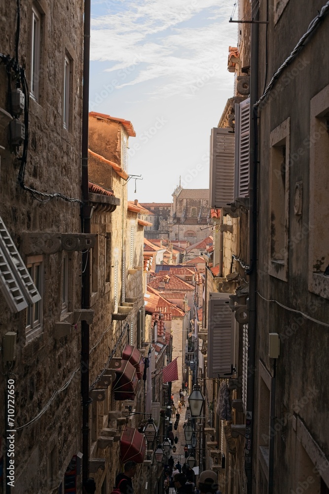 A bustling narrow street winds through the historic limestone buildings of Dubrovnik, Croatia, lined with red roofs, lanterns.
