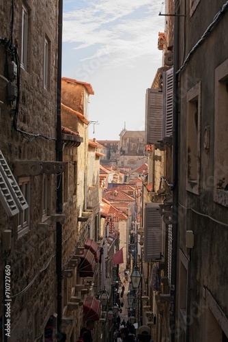 A bustling narrow street winds through the historic limestone buildings of Dubrovnik, Croatia, lined with red roofs, lanterns. © vanzyst